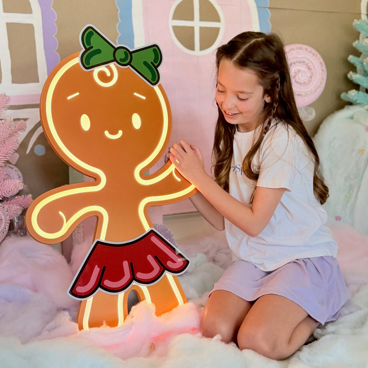 A young girl with long brown hair sits on a fluffy white rug next to a large, glowing gingerbread person decoration. The decoration features a brown body with a green bow and a red skirt, outlined in warm yellow light, with pink glow accents at the base.