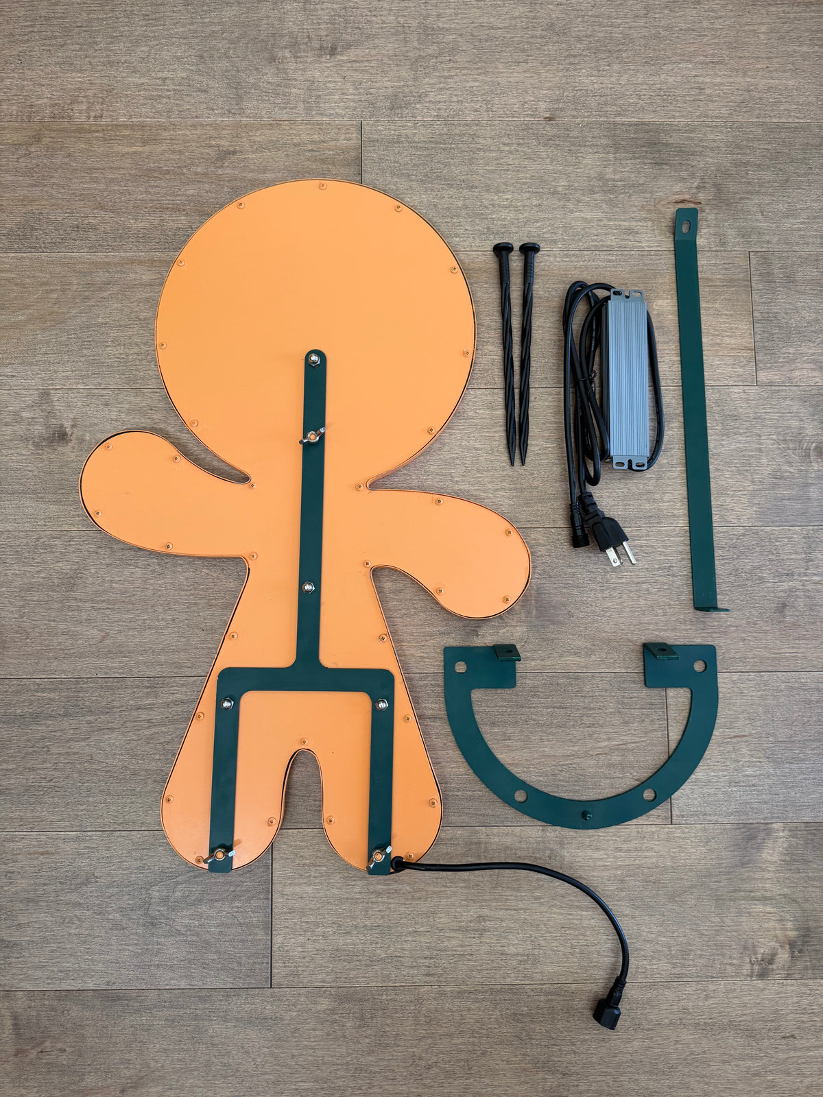 LED gingerbread wall light kit with its mounting hardware, including two nails, a bracket, a strap, and a power adapter, laid out on a gray wood-plank floor.