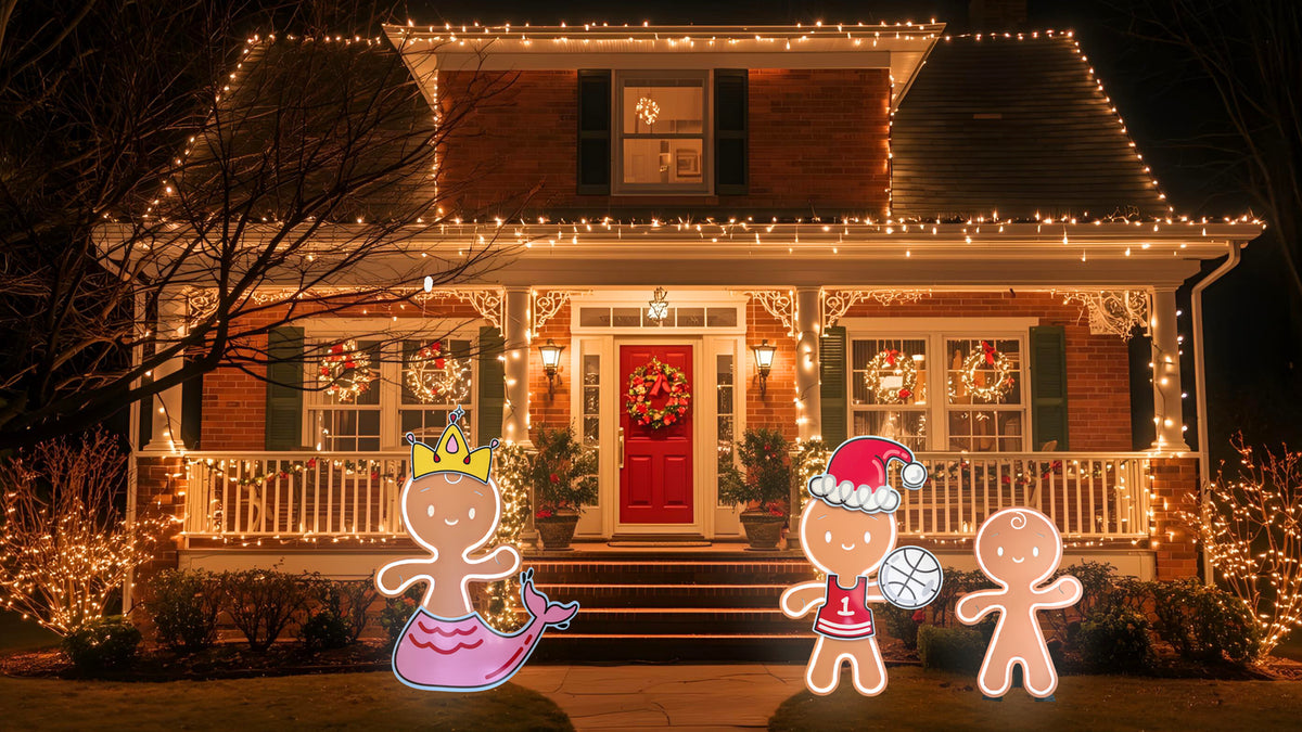 A front yard at night with a decorated house covered in warm white string lights, a red front door with a wreath, and three gingerbread-themed yard signs in the lawn.