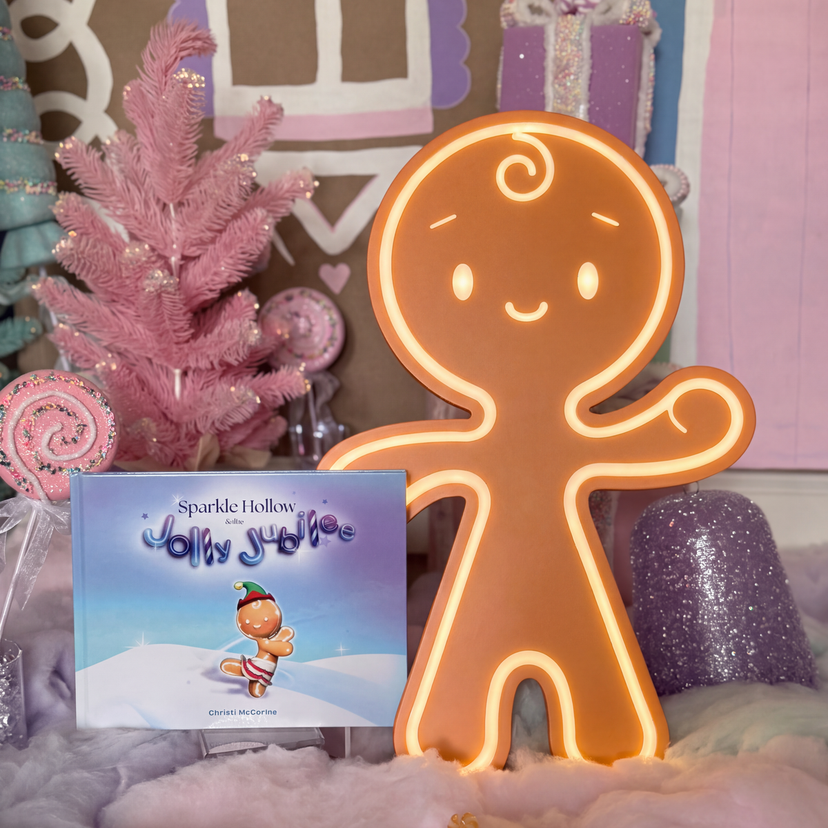 A glowing gingerbread person light stands next to a children's book titled 'Sparkle Hollow Jolly Stroll' on a soft pink and white fluffy surface, with a pink tree and pastel decorations in the background.